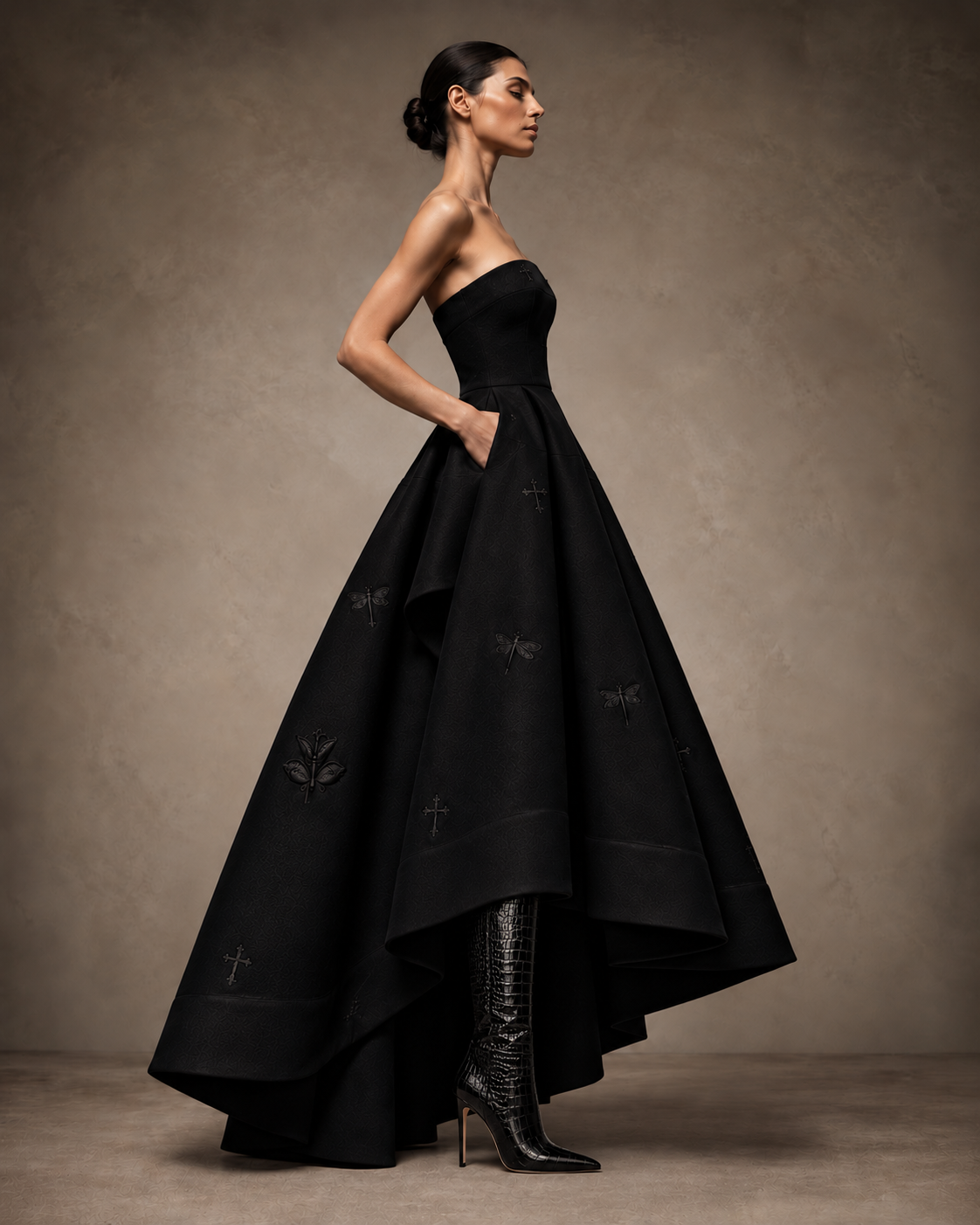 Side view of the black strapless embroidered high-low gown, showing the sculptural skirt, side pocket and floor-length back.